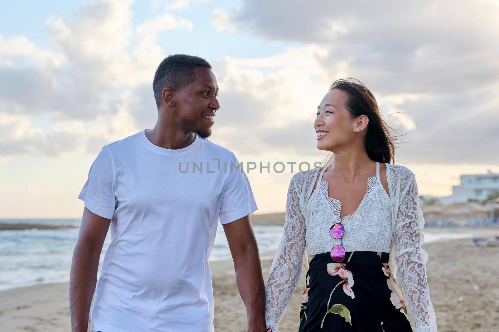 Young happy couple walking on beach holding hands. Multicultural couple in love together on sea shore. Honeymoon, dating, happiness, relationship, vacation, love, romance concept