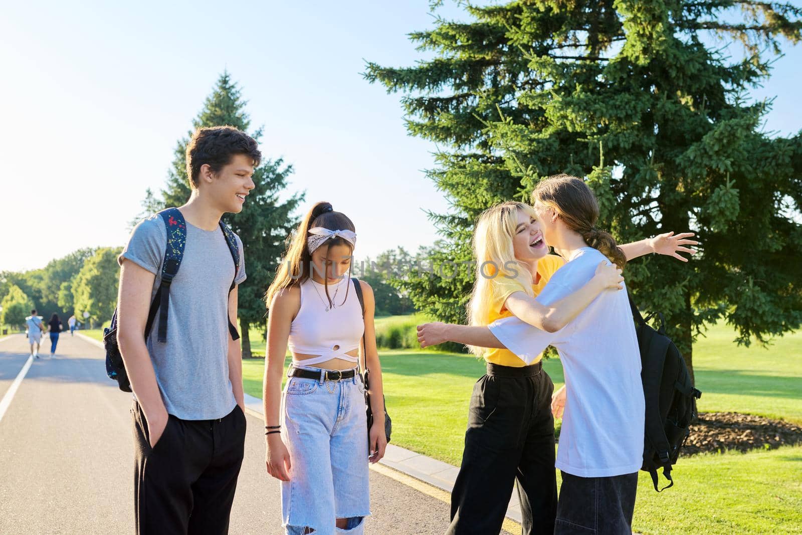 Meeting of smiling teenage friends in sunny summer park. Happy young people greeting each other, hugging, laughing, talking. Friendship, adolescence, communication, emotions, youth concept