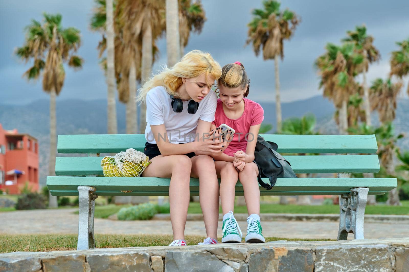 Friendly family, two girls sisters, teenage and preteen look at smartphone. Sitting in park with tropical trees, summer vacation together