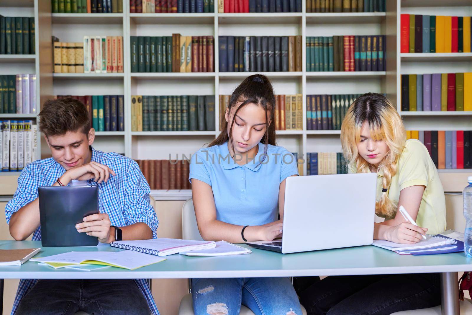 Group of teenage students studying in the school library, writing readers using digital tablet, laptop. Education, knowledge, adolescence, school, college concept