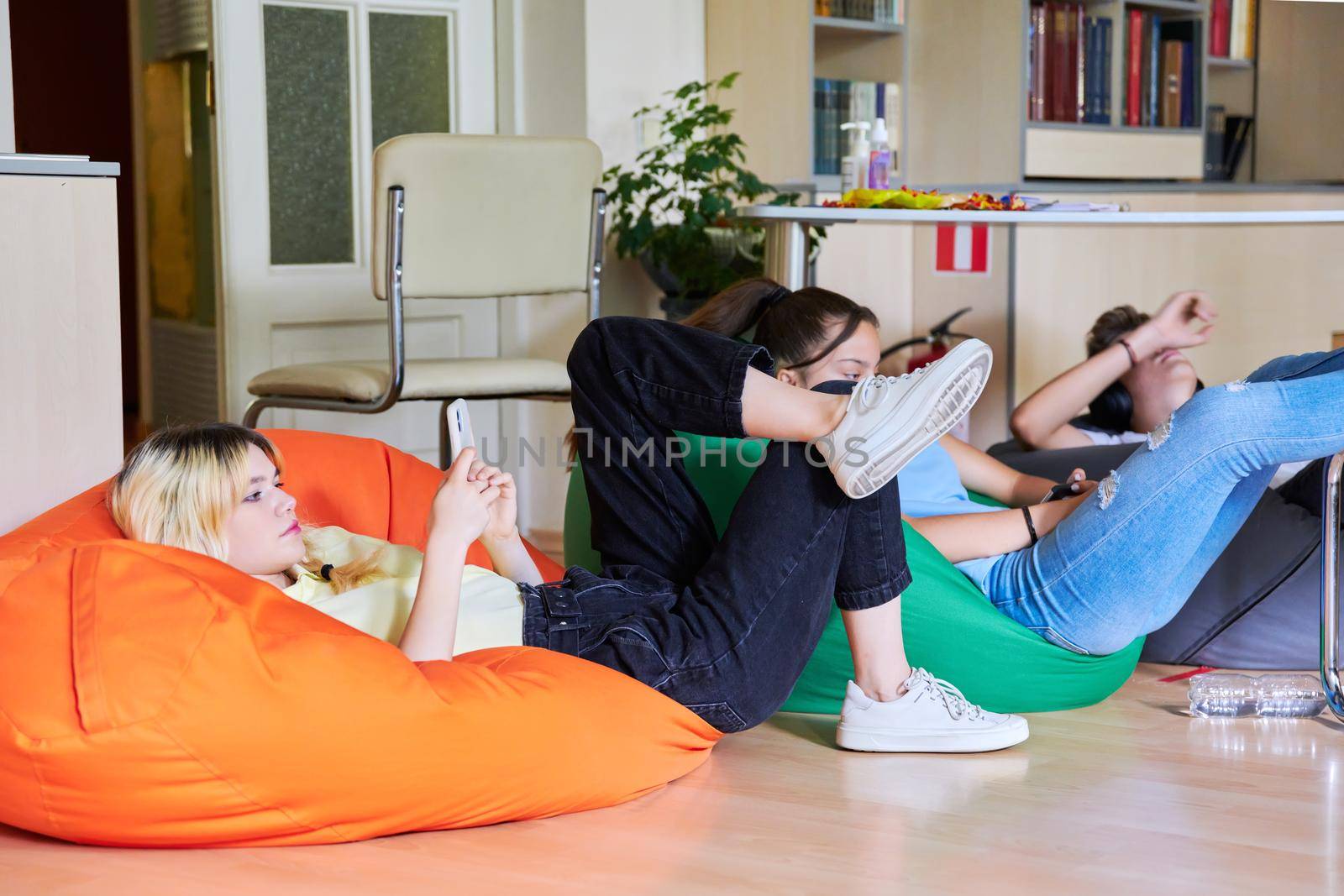 Teenage students resting in the classroom on bean bags on the floor. Adolescence, high school, leisure concept