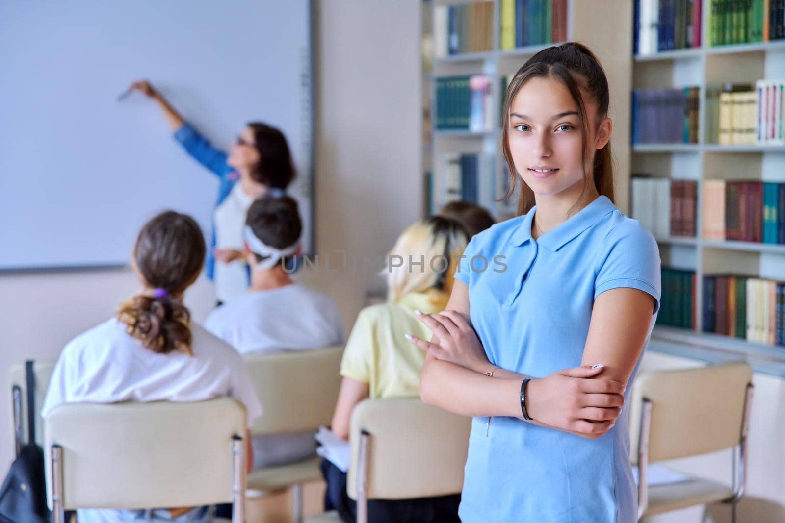 Female teenager student smiling looking at camera, group of pupils and teacher in library. High school, college, knowledge, education, adolescence concept