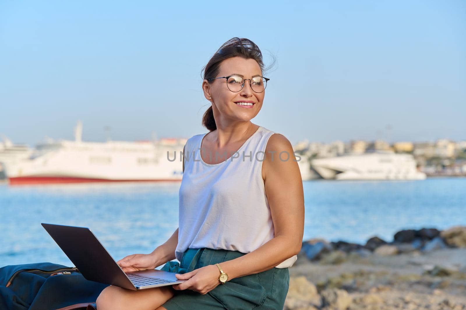 Middle-aged woman tourist with a backpack, traveling on the sea, in the seaport on a bench using a laptop. Technology, vacations, tourism, leisure, active lifestyle, mature people concept