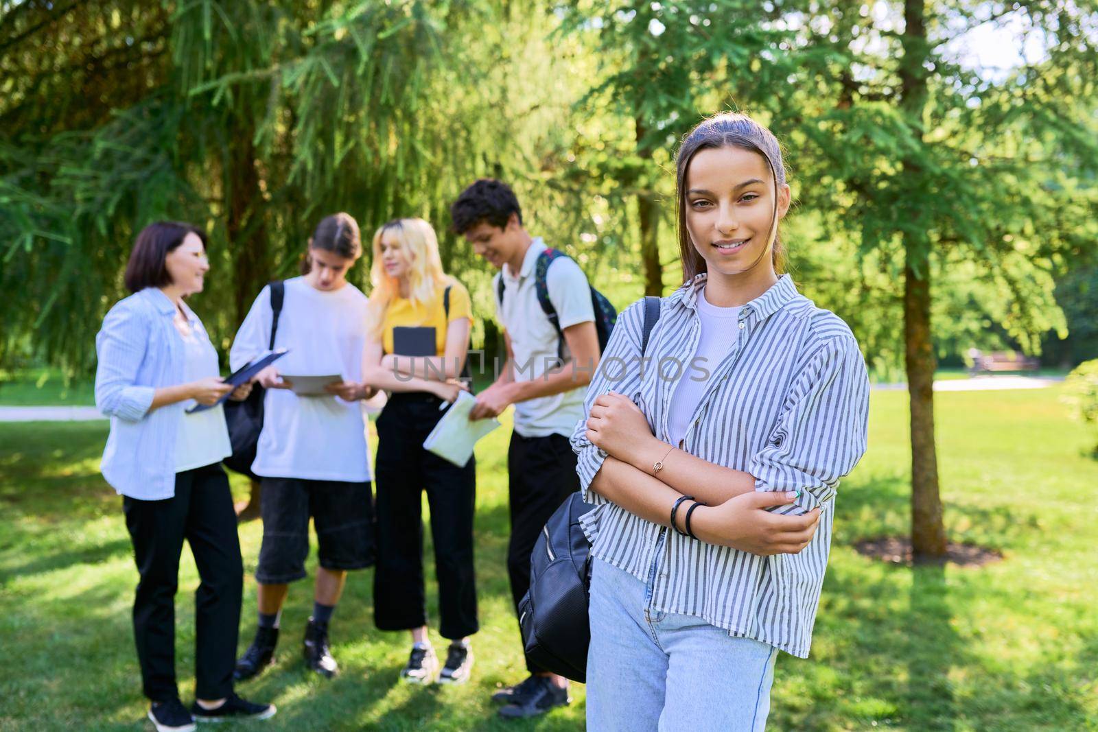 Female student 14, 15 years old with textbooks backpack, group of teenagers talking with teacher in park background. Education, teenage students, adolescence, school concept