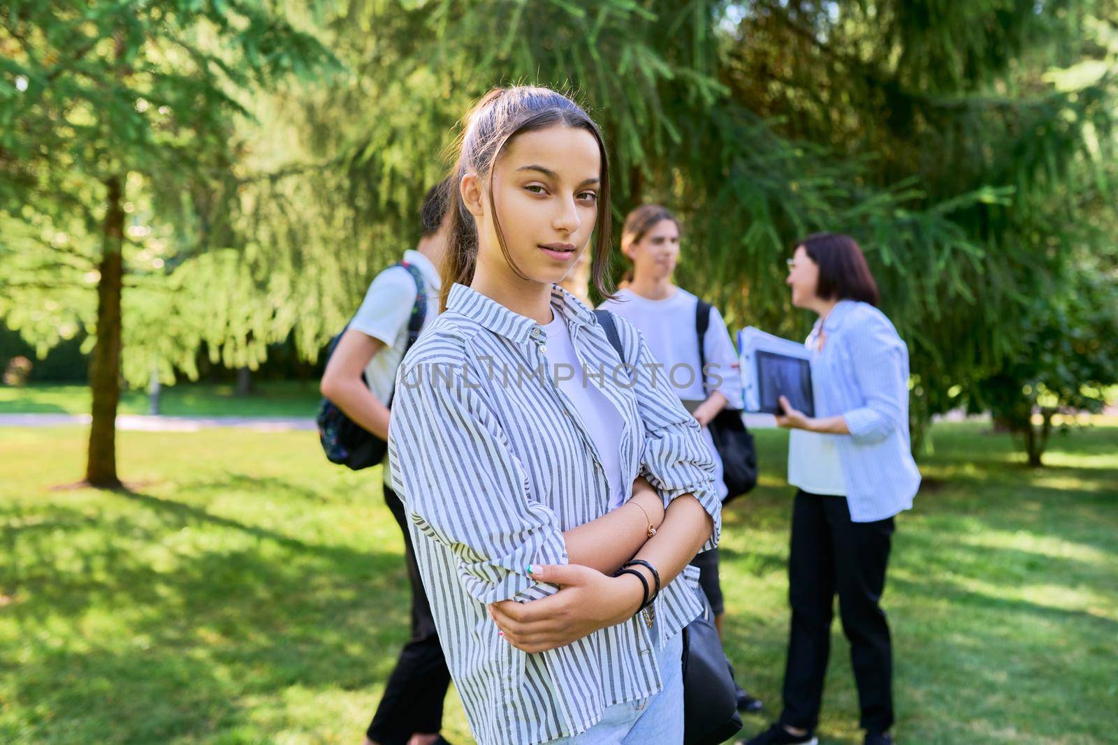 Female student 14, 15 years old with textbooks backpack, group of teenagers talking with teacher in park background. Education, teenage students, adolescence, school concept