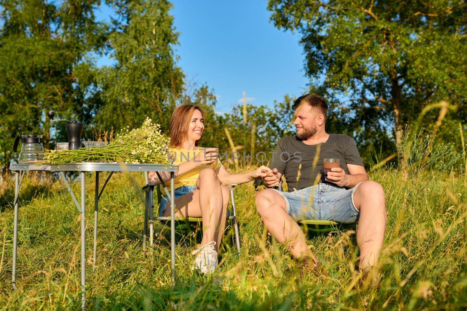 Happy middle aged couple relaxing together outdoor sitting on folding chairs at camping table. Man and woman talking, drinking fresh coffee, enjoying summer nature of wild meadow. People 40s, leisure