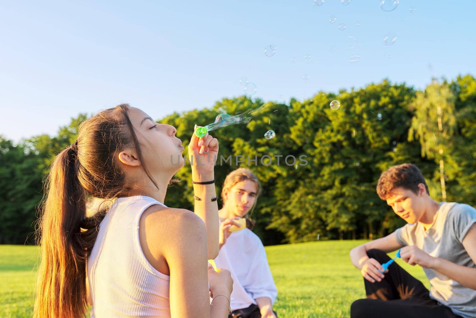 Happy teenage girl 13, 14 years old blowing bubbles. Teenager having fun with friends on lawn in park on sunny day. Fun, summer, vacation, friendship, adolescence concept