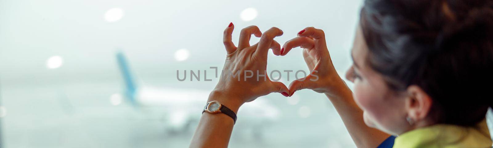 Close up of woman stewardess making heart sign with hands while looking out the window in airport terminal