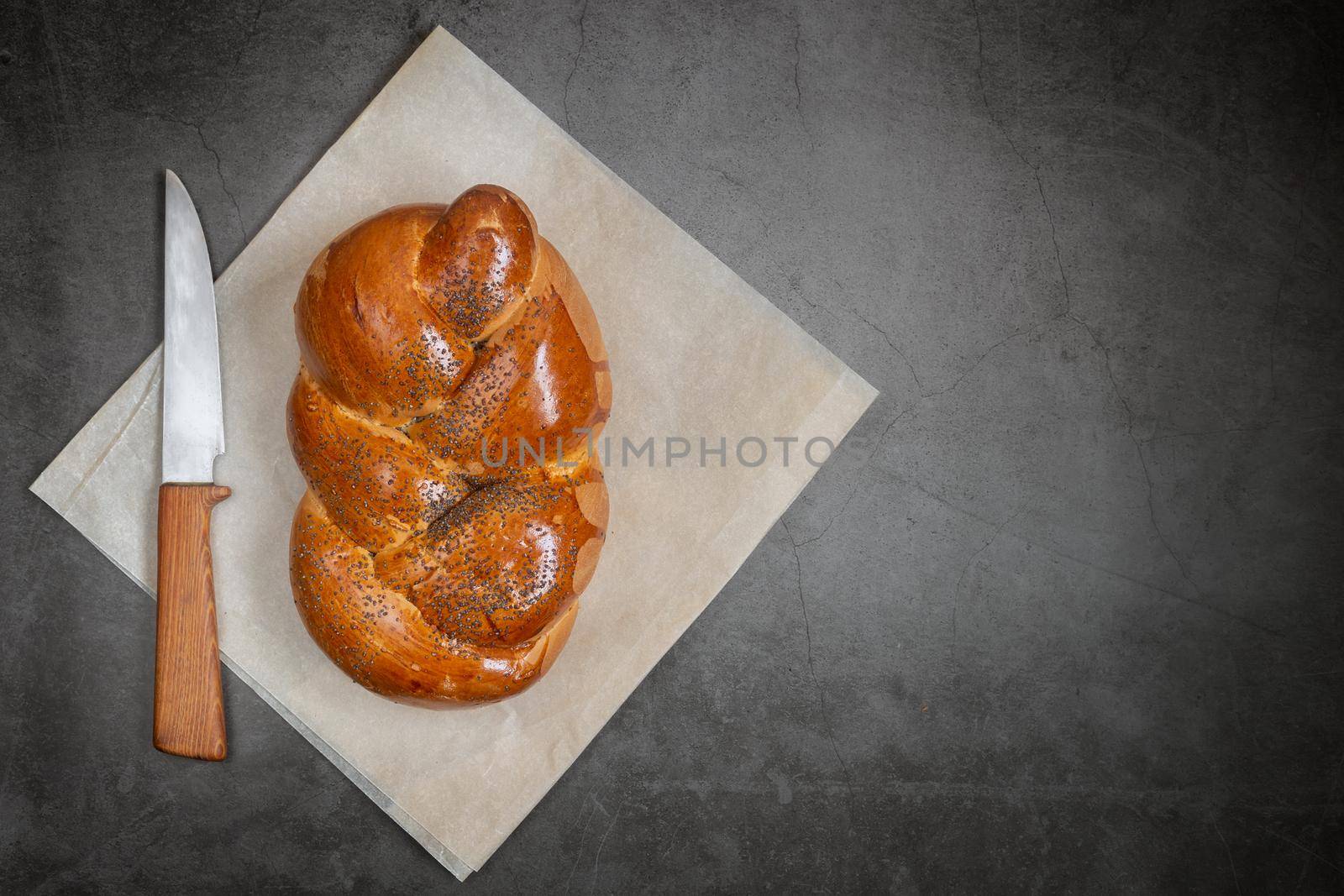 On a linen napkin is a loaf of white bread in the form of a pigtail with poppy seeds and crispy crust and a knife. Presented on a dark background, top view, copy space