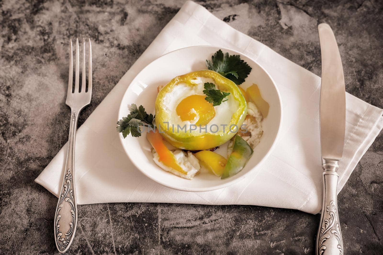 On the table on a plate is an oven-baked bell pepper with an egg inside the pepper. There are Cutlery on a napkin nearby. Top view with space to copy. Flat lay