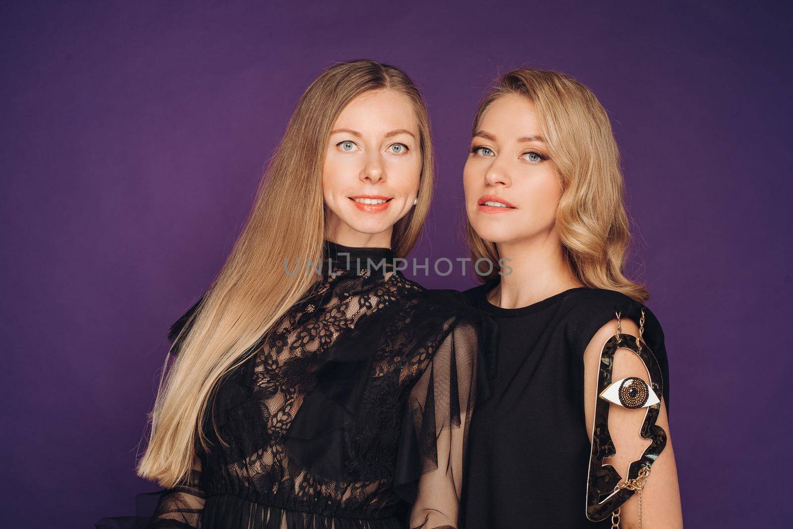 Two sisters pose in the studio on a purple background. 2 beautiful women in black fancy dresses gathered for a party. by StudioLucky