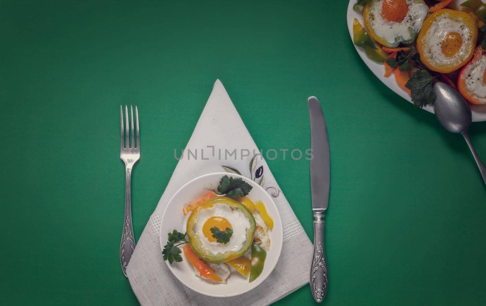 On the table on plates are baked in the oven bell peppers with an egg inside each pepper. There are Cutlery on a napkin nearby. Top view with space to copy. Flat lay