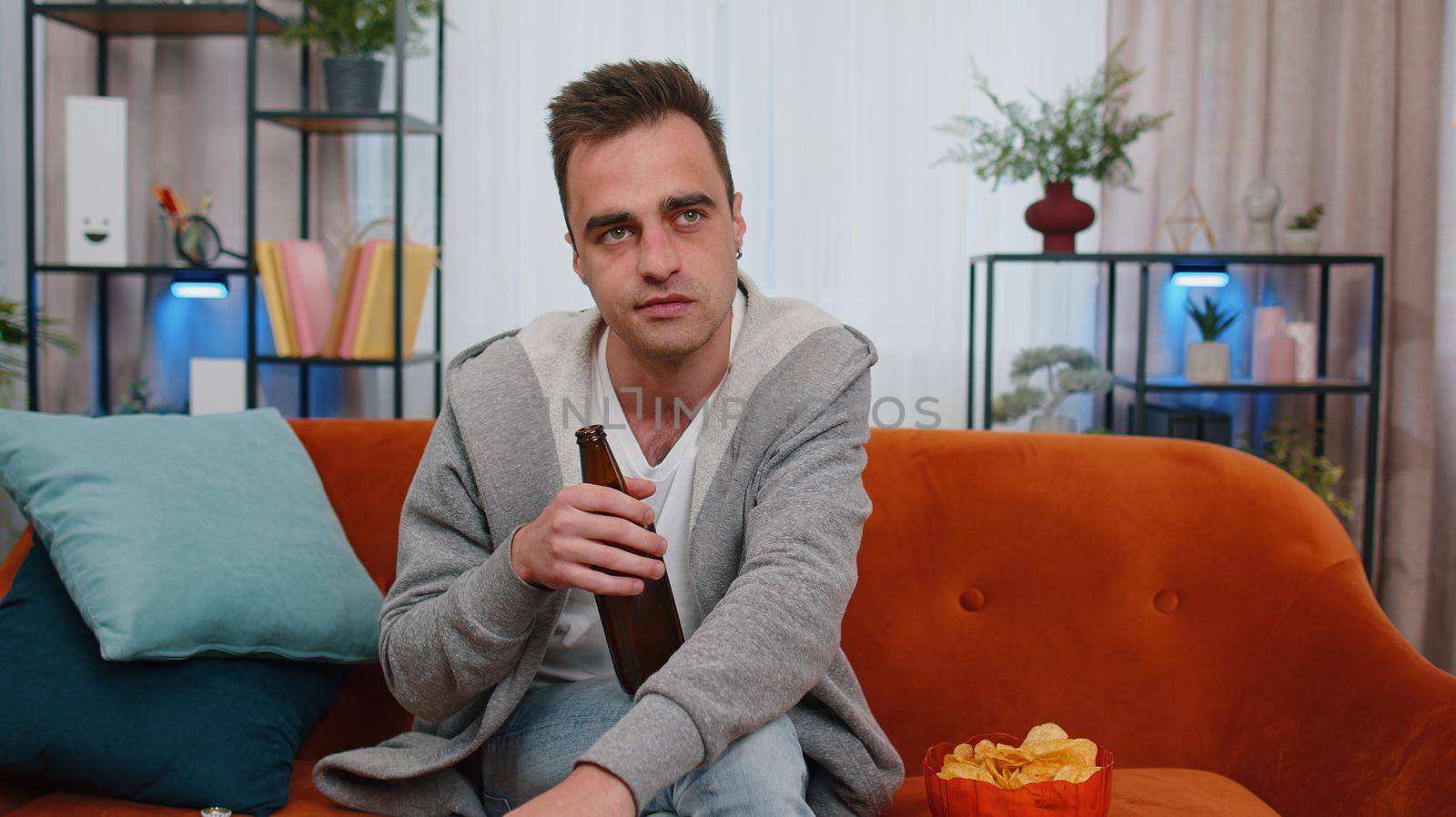 Portrait of man sitting on sofa, eating chips, drinking beer and watching interesting tv serial, football championship game, social media content online at home. Guy enjoying domestic entertainment