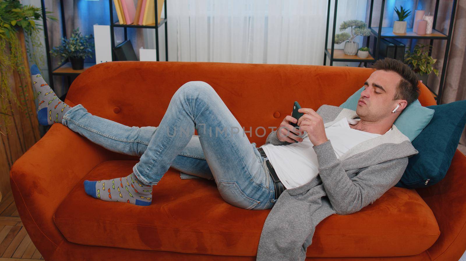 Happy relaxed caucasian man in wireless earphones dancing lying on couch at home choosing listening favorite energetic disco music in smartphone app entertaining indoors. People weekends activities