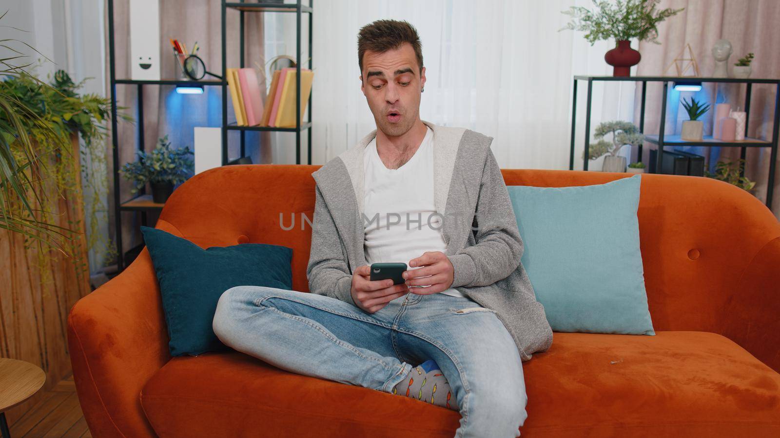 Oh my God Wow. Excited happy joyful winner man use smartphone typing browsing found out great big win good news celebrate victory. Young guy with mobile phone sitting at home in living room on couch