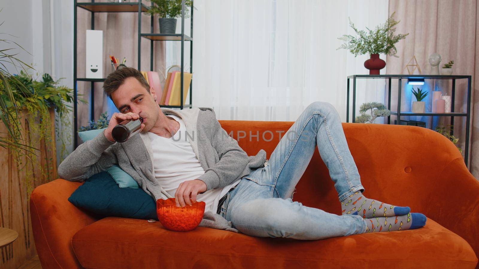 Portrait of man lying on sofa, eating chips, drinking beer and watching interesting tv serial, football championship game, social media content online at home. Guy enjoying domestic entertainment