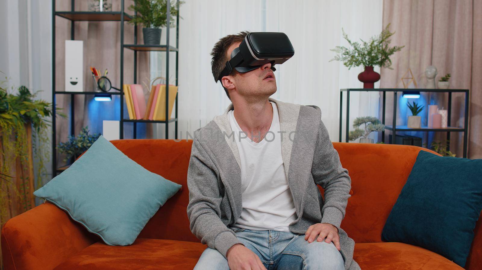 Surprised man use VR headset glasses at home, enjoying 3D 360 video game concept, entertaining, in positive mood, moving hands in air, sitting in living room. Virtual reality, modern gadget device