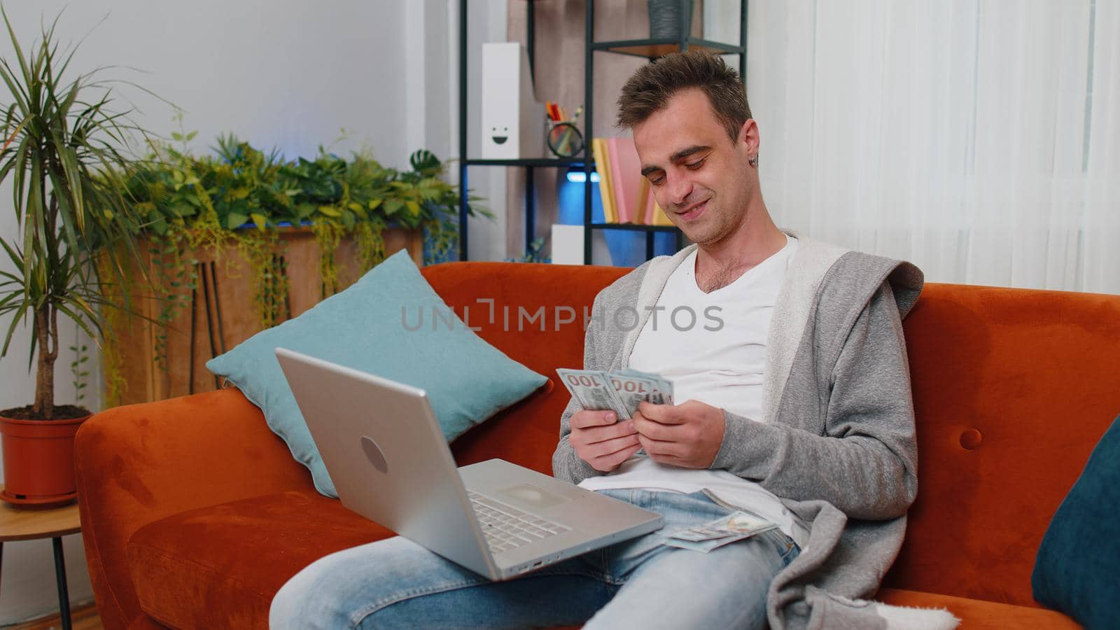 Planning budget. Smiling rich happy young man counting money cash use laptop computer calculate domestic bills at home. Joyful guy satisfied of income earnings, saves money for planned vacation, gifts
