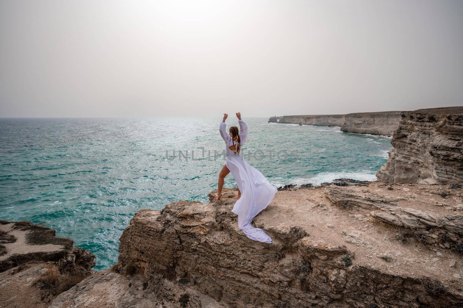 Happy freedom woman on the beach enjoying and posing in white dress over the sea. View of a girl in a fluttering white dress in the wind. Holidays, holidays at sea