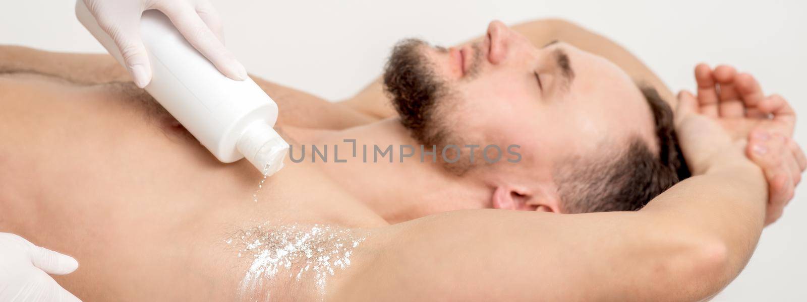 Hand of master depilation pouring talcum powder on armpit of young man before depilation procedure.