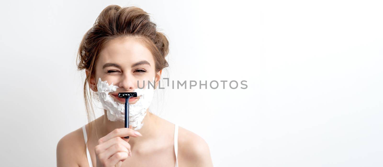 Beautiful young caucasian woman shaving her face by razor on white background. Pretty woman with shaving foam on her face