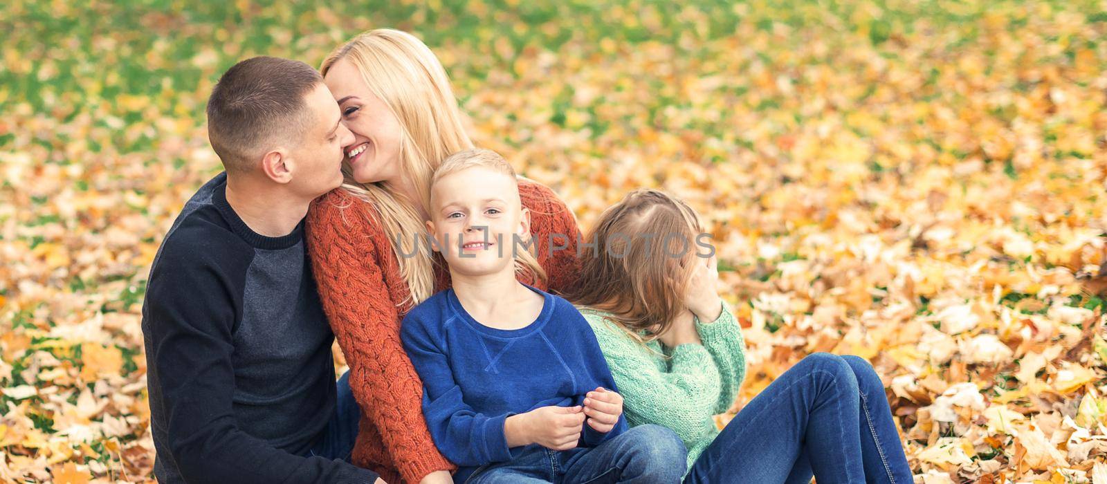 Portrait of young family sitting in autumn leaves. Parents kissing and sitting with children in the autumn park