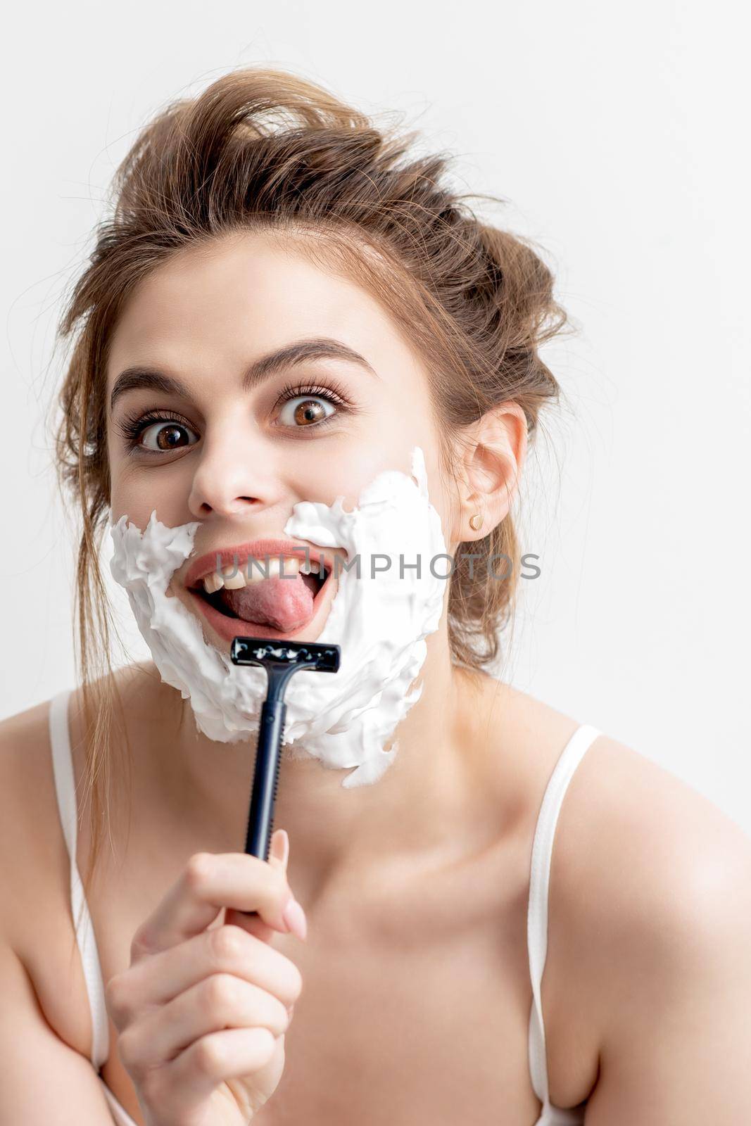 Beautiful young caucasian woman shaving her face by razor on white background. Pretty woman with shaving foam on her face