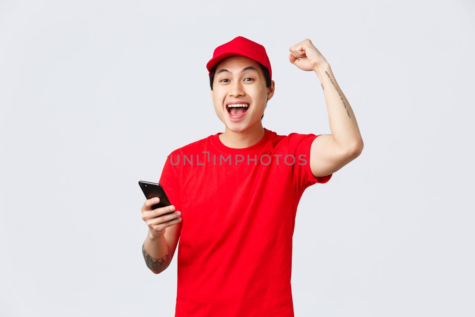 Happy, cheerful asian delivery guy in red uniform cap and t-shirt of carrier service, holding smartphone, reading good news, chanting with fist pump, yell yes celebrate victory or achivement.