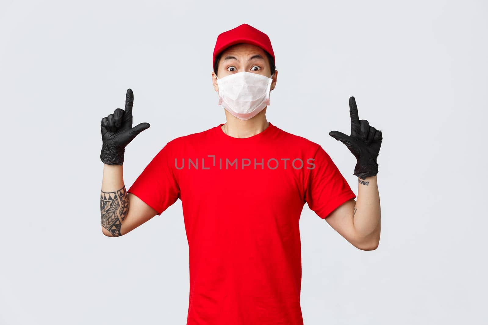 Surprised young asian delivery guy in company uniform, red t-shirt and cap, staring camera impressed and shocked, pointing fingers up, wear protective gloves and medical mask while at work.