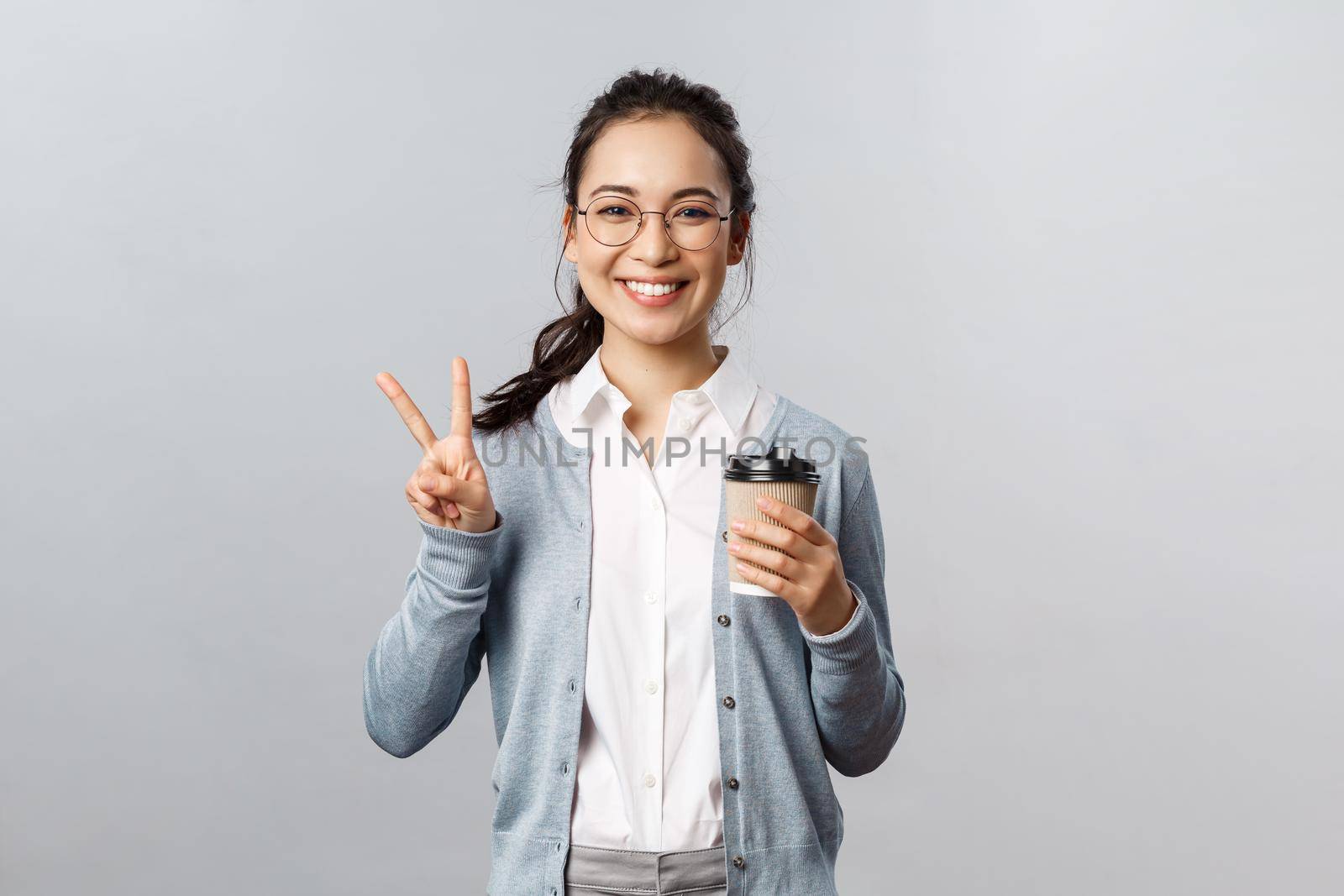 Office lifestyle, business and people concept. Optimistic, friendly-looking asian girl show peace sign, smiling kawaii, enjoying morning fresh cup coffee from local cafe, stand grey background by Benzoix