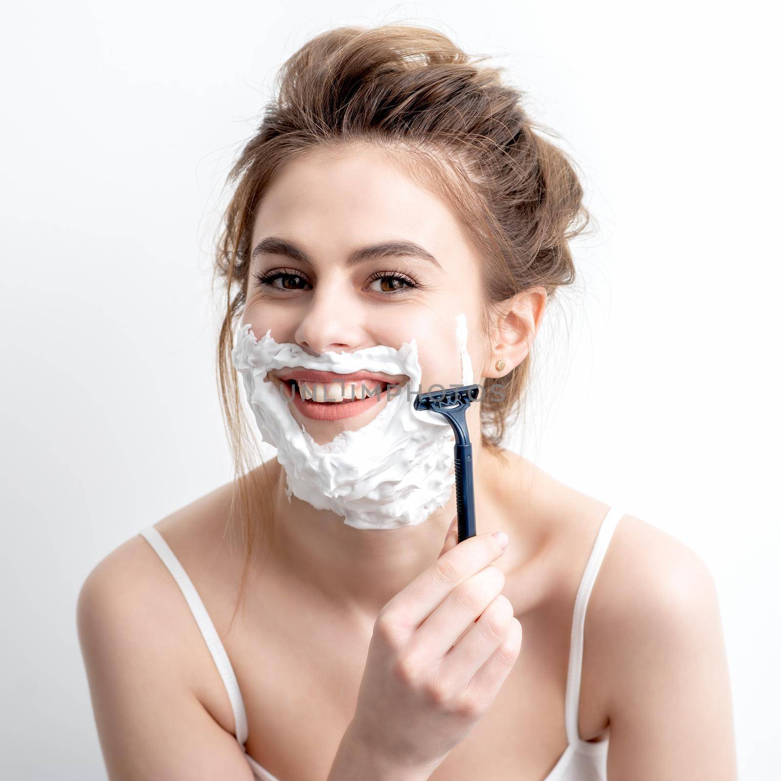 Beautiful young caucasian woman shaving her face by razor on white background. Pretty woman with shaving foam on her face