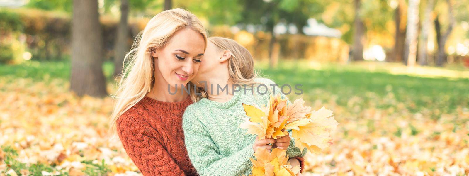 Happy young caucasian mother and little daughter holding autumn yellow leaves sitting and kissing at the park