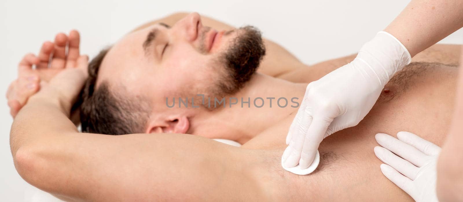 Hands of depilation master in white rubber gloves wiping male armpit with a cotton pad