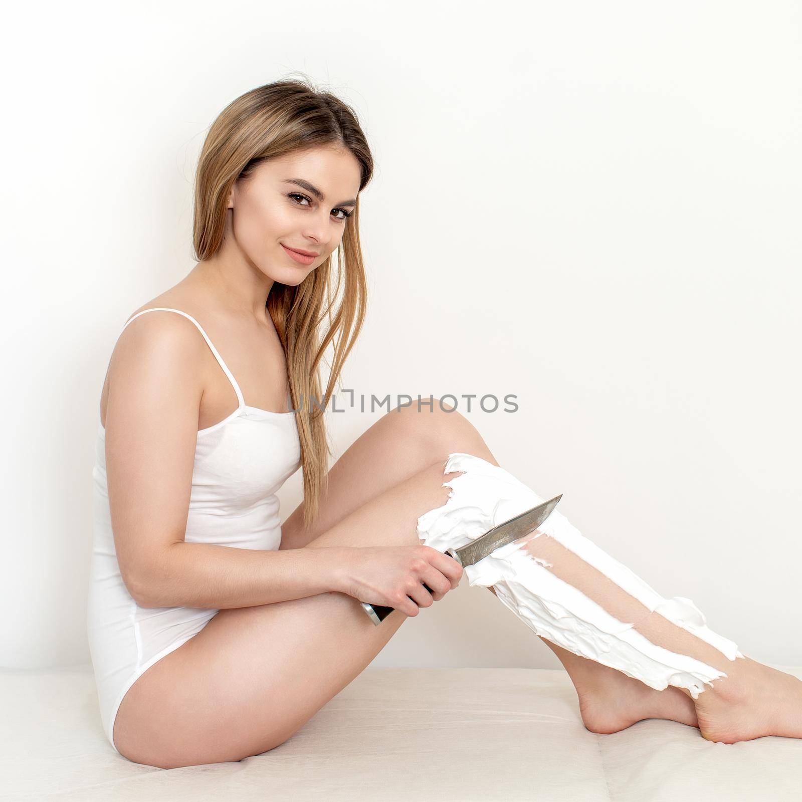 Beautiful young caucasian woman shaves her legs with a knife on a white background