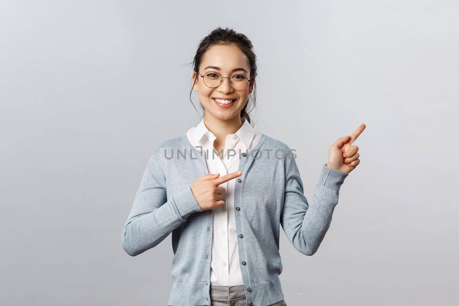 Education, studying and online classes concept. Cheerful asian woman, teacher pointing fingers right at presentation or chart, smiling friendly, helping find way, showing promo, grey background by Benzoix