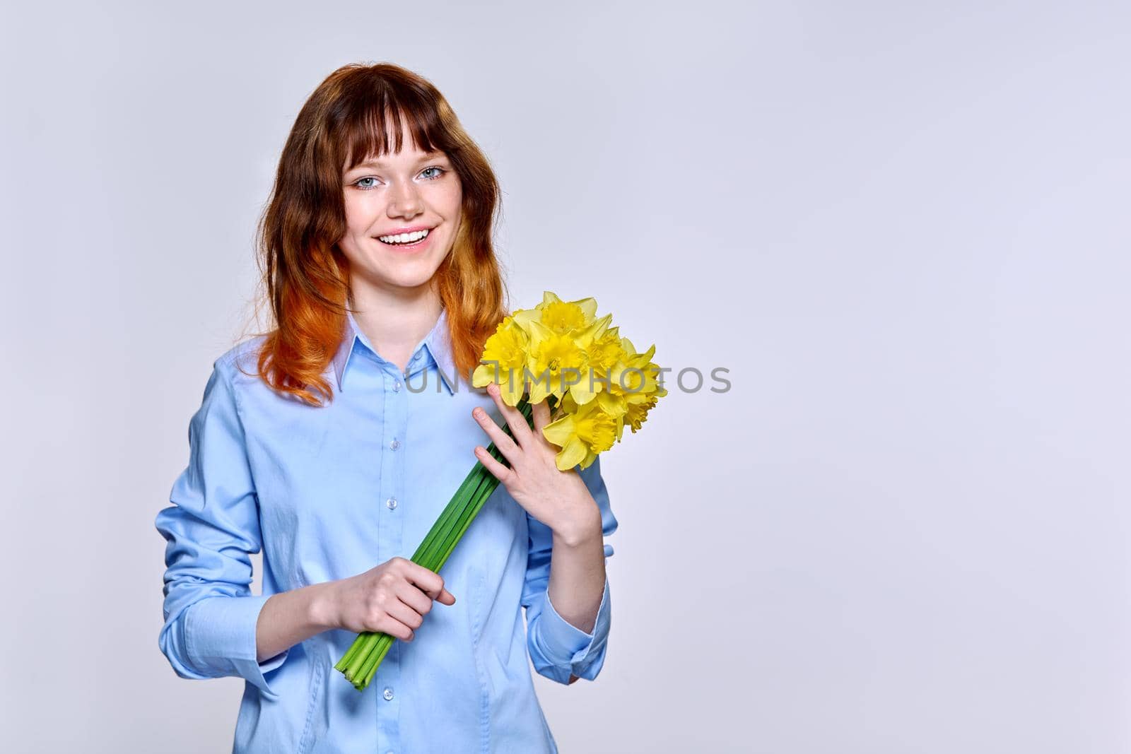 Portrait of young woman with bouquet of yellow flowers looking at camera, copy space, light background. Beautiful happy red-haired teenage female in blue shirt. Happiness, joy, holiday, beauty, youth