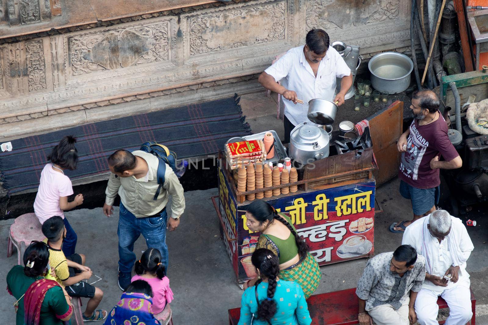 aerial drone shot of road side tea seller in white shirt kurta with small stall preparing tea on a stove and old utensils by putting in milk, leaves, sugar and more as the crowds move around him in the busy city by Shalinimathur