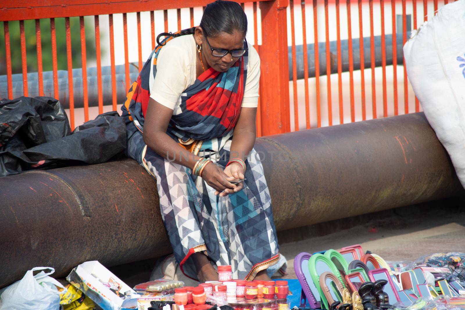 Old streetside shopkeeper making handmade bead necklaces for sale sitting under a plastic sheet, haridwar houses a lot of these old destitute people working to make ends meet by Shalinimathur