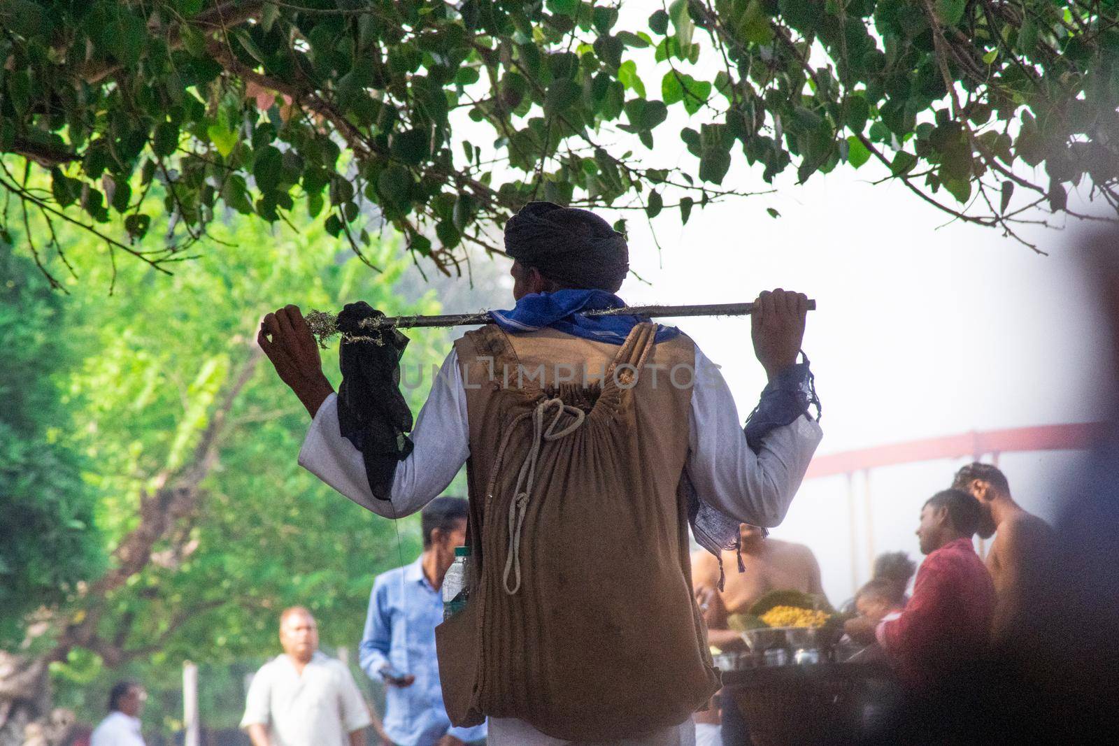 Haridwar, Uttarakhand, India - circa 2021 : bnajara wanderer with stick and backpack with turban walking along river side ghat stopping to pick leaves from a tree as the crowd around him goes about their business