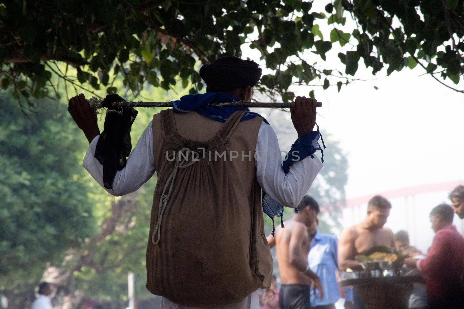 Haridwar, Uttarakhand, India - circa 2021 : bnajara wanderer with stick and backpack with turban walking along river side ghat stopping to pick leaves from a tree as the crowd around him goes about their business