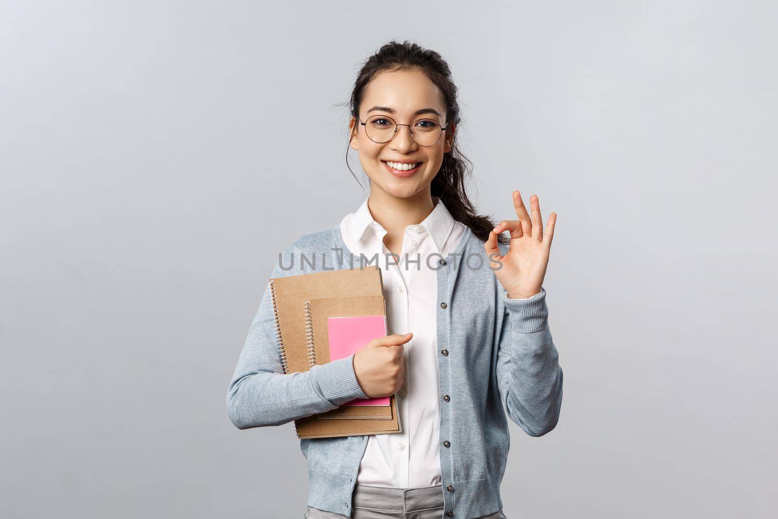 Education, teachers, university and schools concept. Excellent school, join our online lesson. Portrait of satisfied young female student in glasses, show okay sign and hold notebooks with homework.