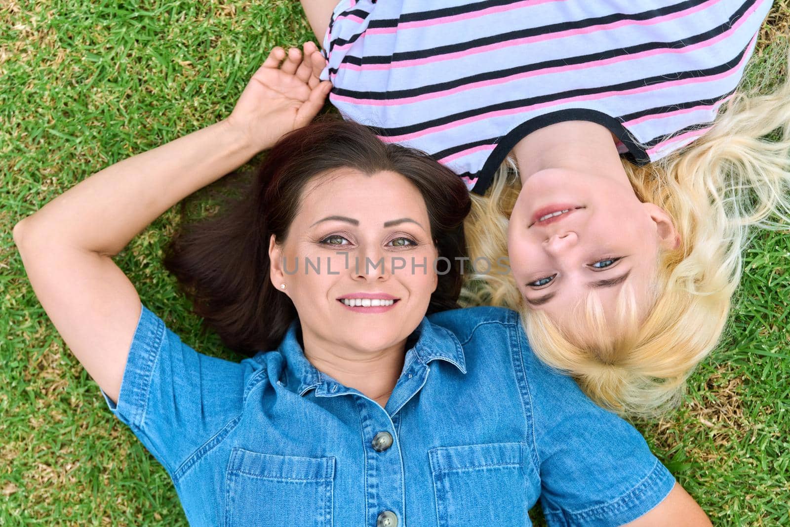 Closeup face of happy mother and teenage daughter looking at camera, mom and girl lying on grass top view. Motherhood, family, adolescence, relationships, people concept