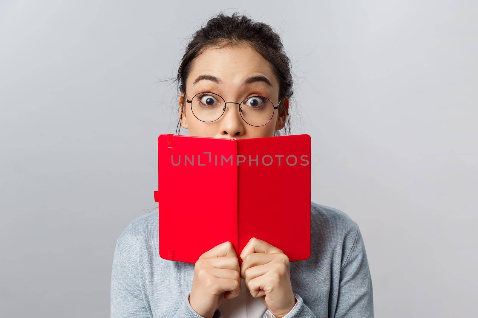 Education, university and people concept. Close-up portrait of surprised and amused young asian girl, widen eyes at amazing interesting story, cover mouth behind red planner, stand grey background by Benzoix