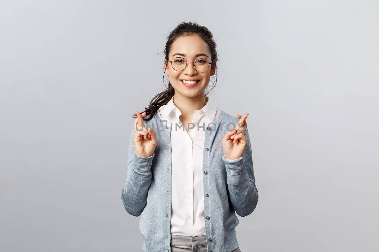 Emotions, people and lifestyle concept. Optimistic happy, smiling asian girl having faith all be alright, cross fingers good luck, hopeful anticipating positive results, pleading over grey background.