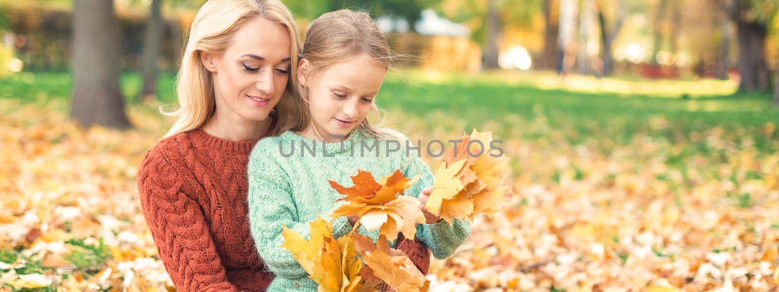 Happy young caucasian woman and little girl holding autumn yellow leaves sitting at the park