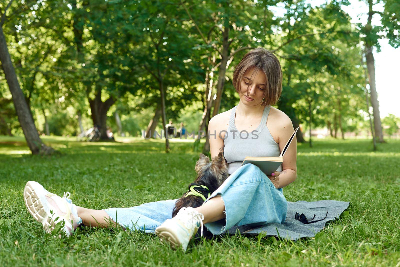 A young woman reads a book in nature next to her small dog Yorshir Terrier.