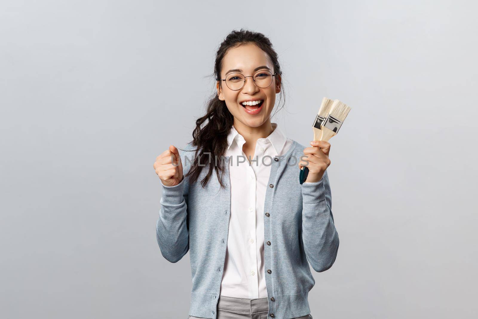 Creativity, repairs and overhaul concept. Happy cheerful asian female encourage roommate start repairment works together, holding two painting brushes and smiling, want change wall color in room by Benzoix