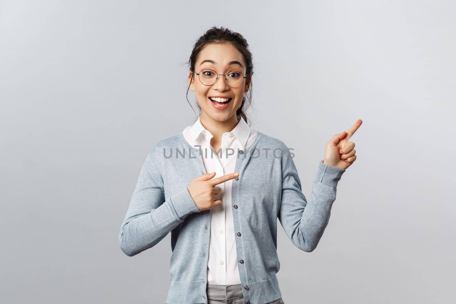 Upbeat excited mixed-race girl in glasses, asian woman teacher pointing fingers right, smiling broadly, found way to stay in touch with students during lockdown quarantine, standing grey background.