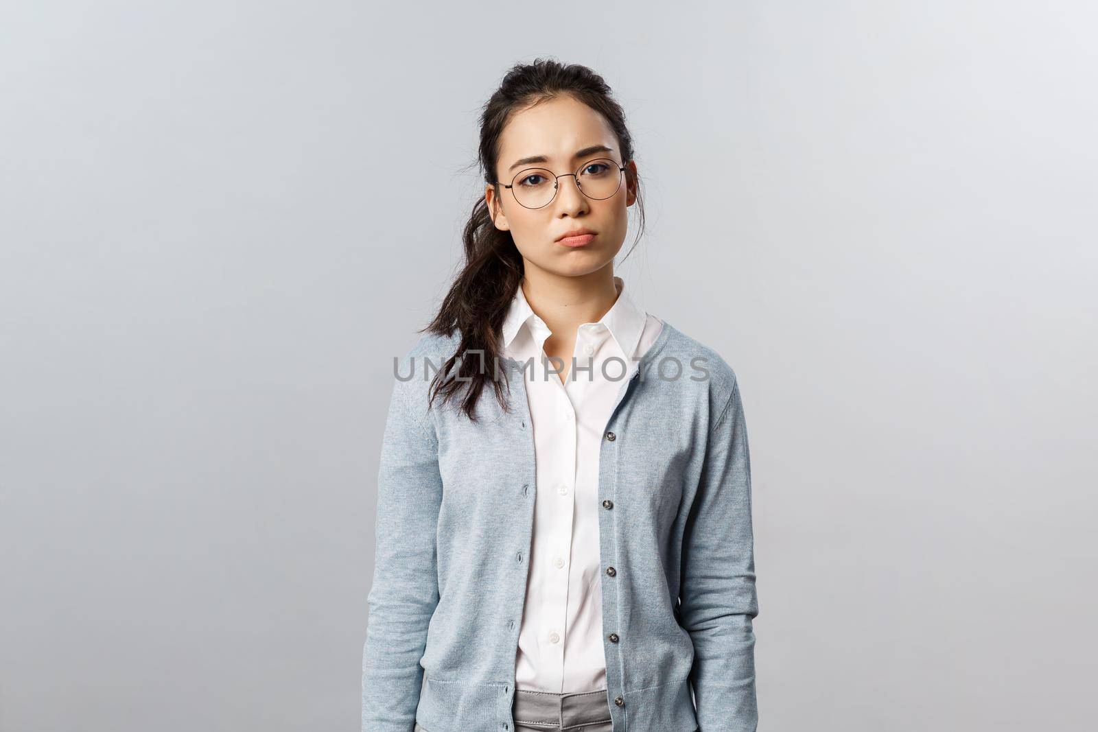 Gloomy and indifferent, lonely asian woman in glasses, feel uneasy and tired look camera drained, feel overworking and displeased, sighing with regret, standing upset over grey background.