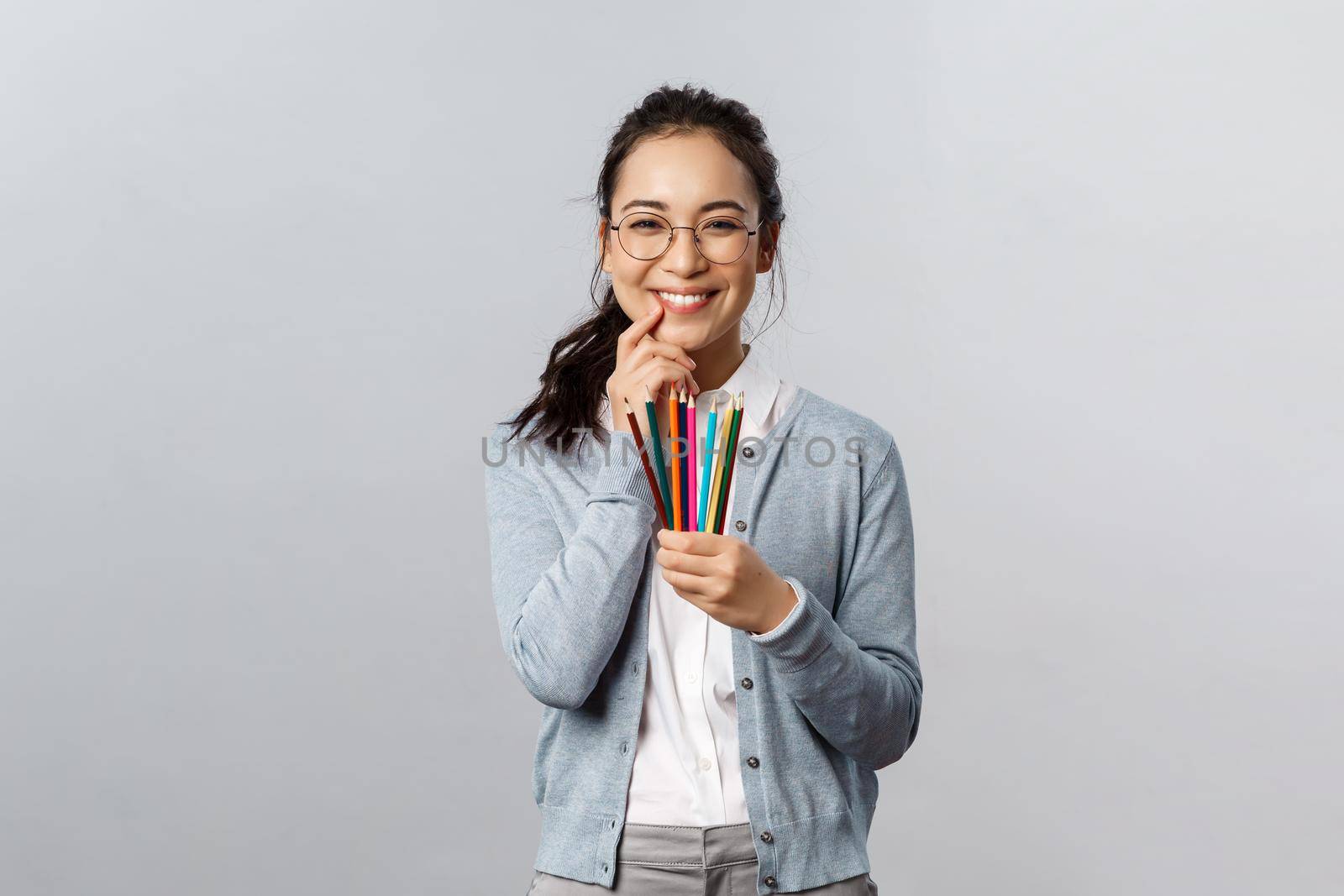 Hobbies, creativity and art concept. Silly happy smiling asian woman, artist in glasses giggle as holding colored pencils, have interesting idea for her next artwork, drawing picture, grey background.